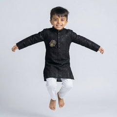 Young boy wearing a black embroidered Lucknowi kurta with white pajamas and white loafers, standing against a white background.