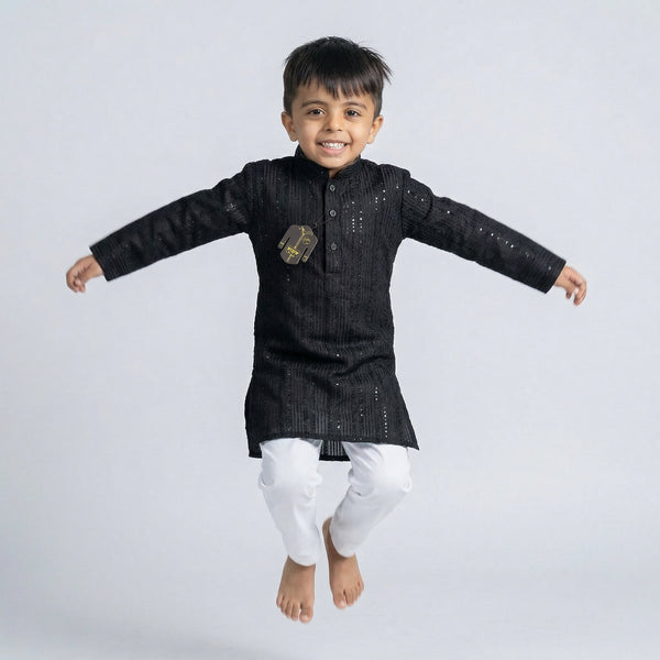 Young boy wearing a black embroidered Lucknowi kurta with white pajamas and white loafers, standing against a white background.