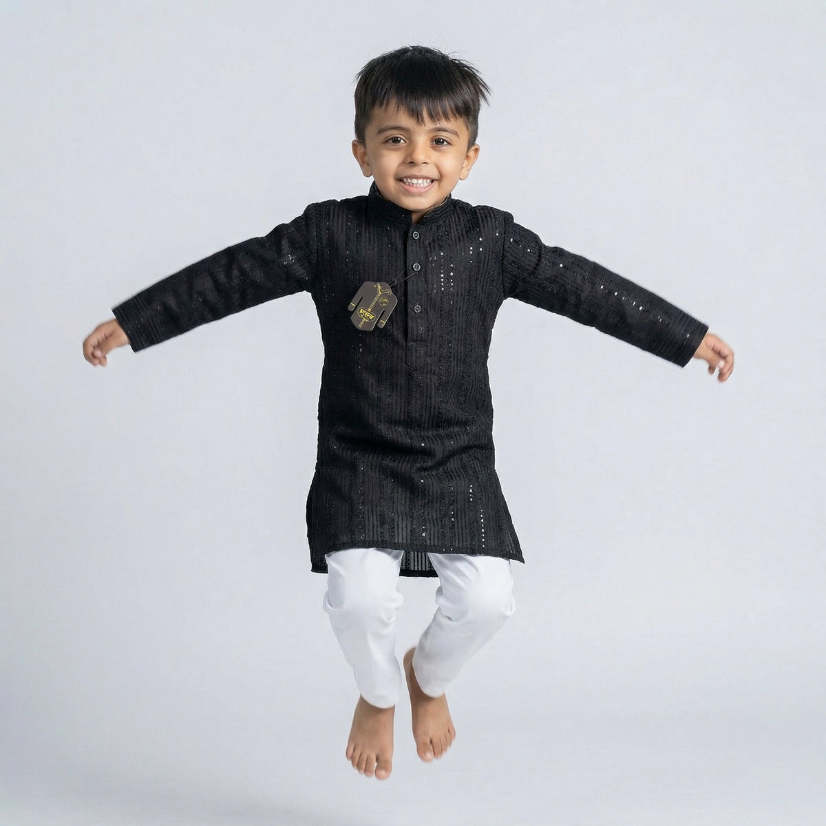 Young boy wearing a black embroidered Lucknowi kurta with white pajamas and white loafers, standing against a white background.