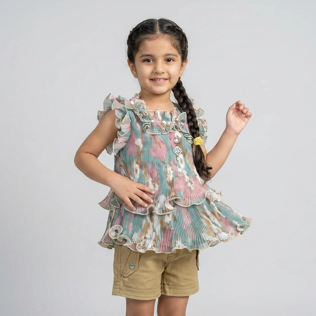A young girl with braided hair wearing a tiered ruffle sleeveless top with a pastel floral print in green, pink, and beige, paired with khaki shorts, against a light gray studio background.