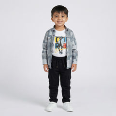 Young boy wearing a grey printed shirt over a white graphic t-shirt with black cargo pants and white sneakers against a light studio background