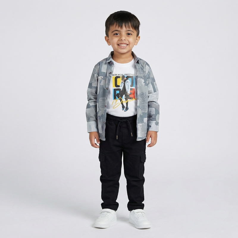 Young boy wearing a grey printed shirt over a white graphic t-shirt with black cargo pants and white sneakers against a light studio background