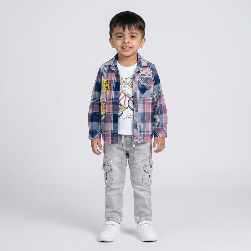 Young boy wearing a pink and blue checked shirt over a white graphic t-shirt with light grey cargo jeans and white sneakers against a light studio background