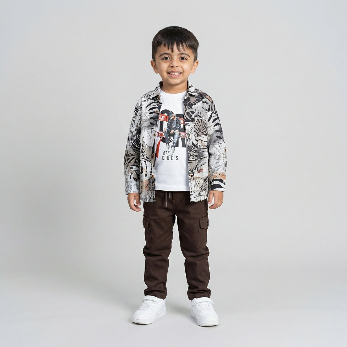 Young boy wearing a tropical print shirt over a white graphic t-shirt with brown cargo pants and white sneakers against a light studio background