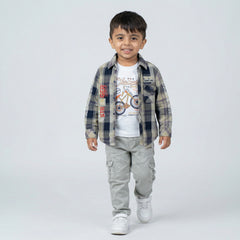 Young boy wearing a grey and blue checked shirt over a white graphic t-shirt with light grey cargo jeans and white sneakers against a light studio background