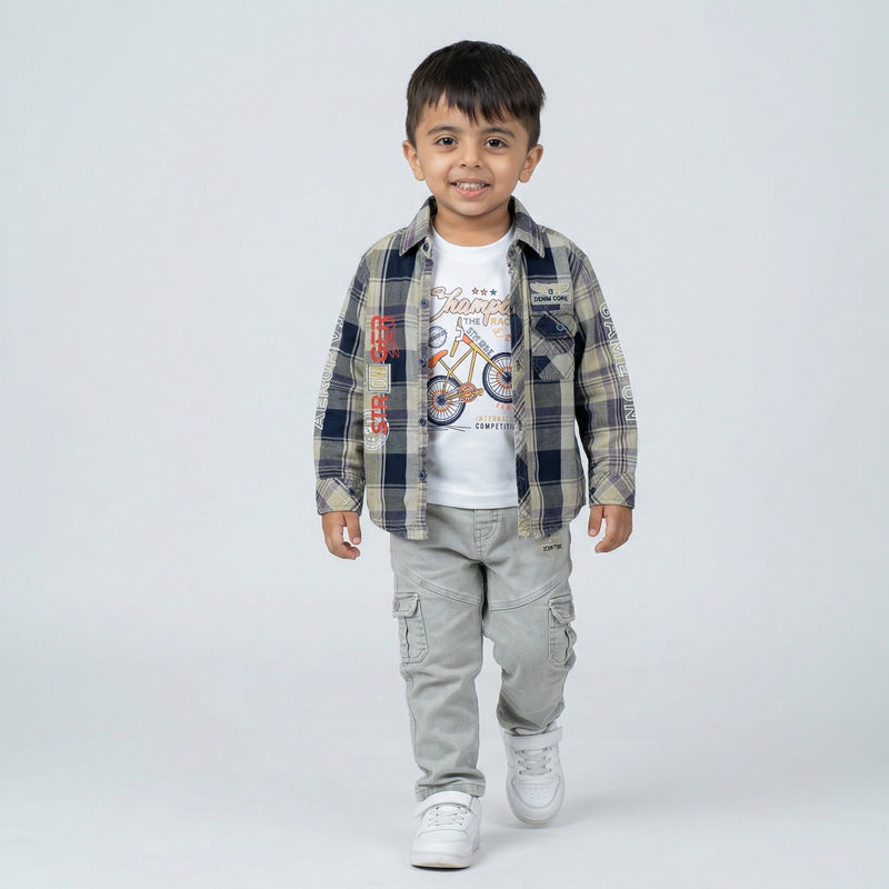 Young boy wearing a grey and blue checked shirt over a white graphic t-shirt with light grey cargo jeans and white sneakers against a light studio background