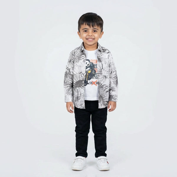 Young boy smiling in a printed casual shirt, white graphic T-shirt, black pants, and white sneakers, standing against a plain light background.