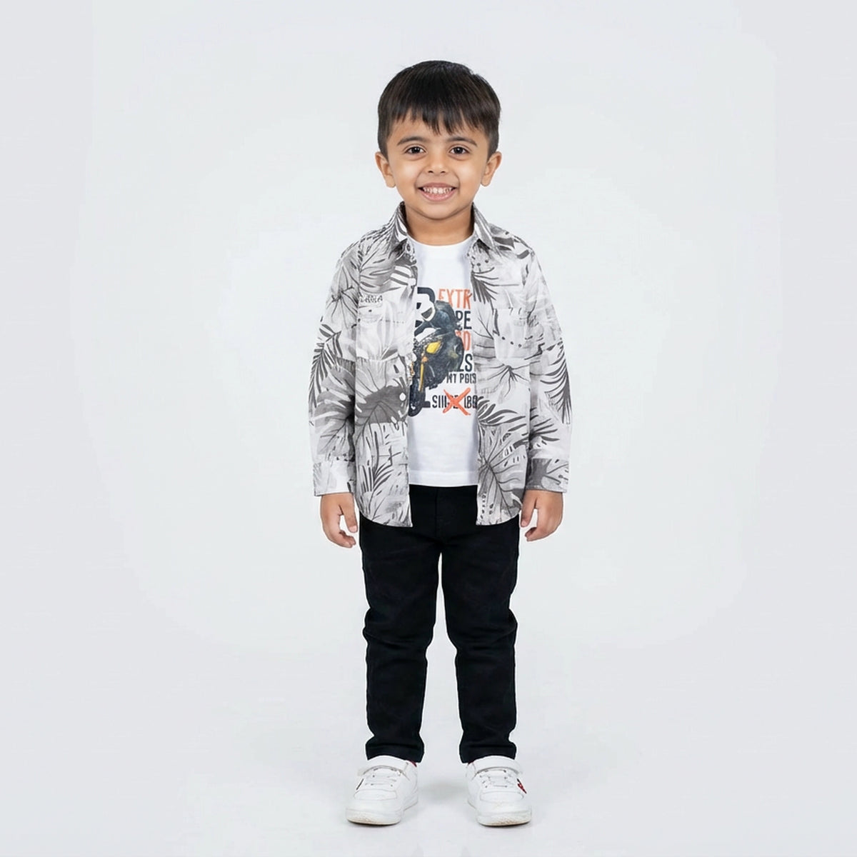 Young boy smiling in a printed casual shirt, white graphic T-shirt, black pants, and white sneakers, standing against a plain light background.