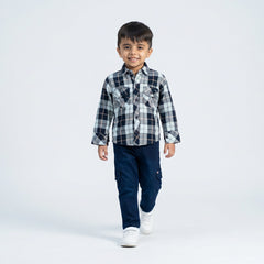 Young boy smiling in a blue and grey checked casual shirt, blue cargo jeans, and white sneakers, standing against a plain light background.