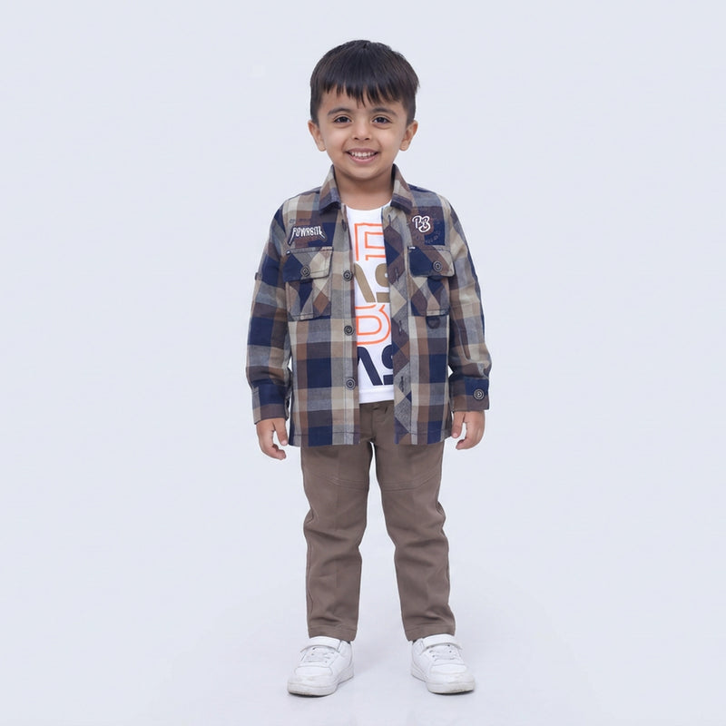 Young boy smiling in a brown checked casual shirt over a white graphic T-shirt, brown pants, and white sneakers, standing against a plain light background.