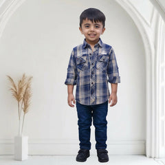 Child wearing a blue plaid shirt and jeans standing in a white room with a plant in the background.