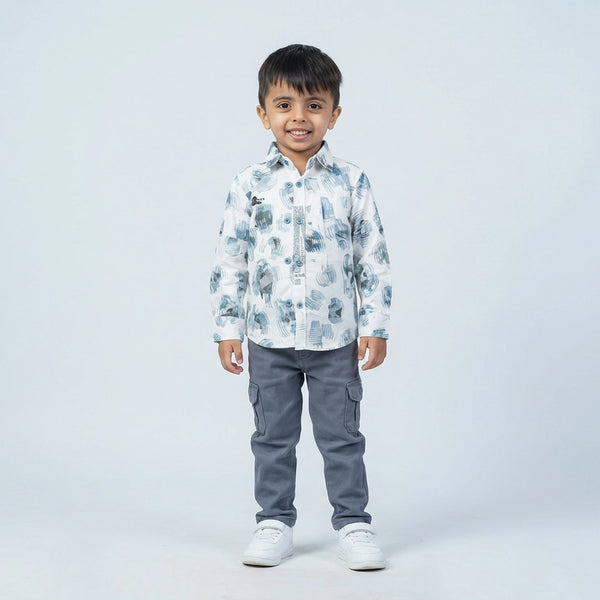 Young boy wearing a white printed full sleeve shirt, grey cargo pants, and white sneakers, standing and smiling against a plain light background.