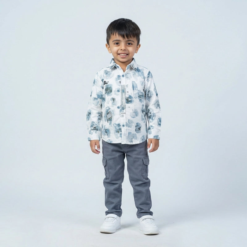 Young boy wearing a white printed full sleeve shirt, grey cargo pants, and white sneakers, standing and smiling against a plain light background.