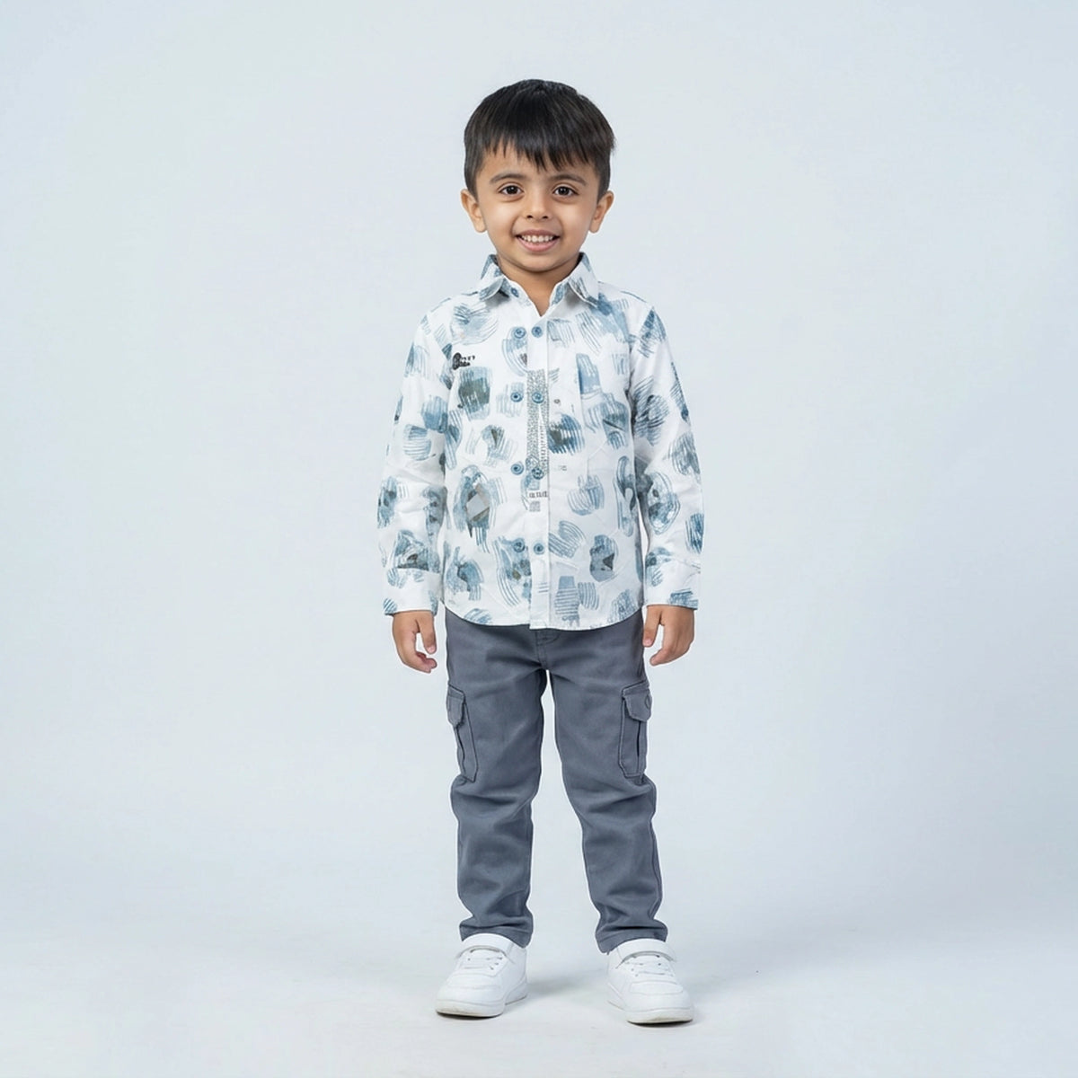 Young boy wearing a white printed full sleeve shirt, grey cargo pants, and white sneakers, standing and smiling against a plain light background.