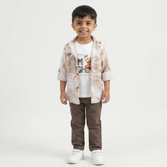 A toddler boy stands smiling in a light printed overshirt, white graphic T-shirt, brown pants, and white sneakers against a plain studio background.