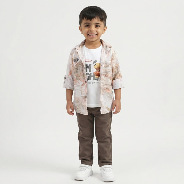 A toddler boy stands smiling in a light printed overshirt, white graphic T-shirt, brown pants, and white sneakers against a plain studio background.