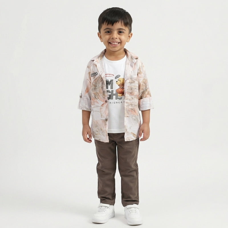 A toddler boy stands smiling in a light printed overshirt, white graphic T-shirt, brown pants, and white sneakers against a plain studio background.