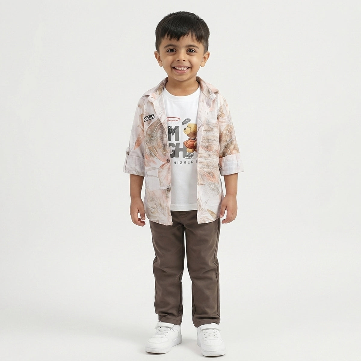 A toddler boy stands smiling in a light printed overshirt, white graphic T-shirt, brown pants, and white sneakers against a plain studio background.