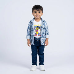 Young boy smiling in a blue printed shirt, cartoon graphic T-shirt, blue jeans, belt, and white sneakers, standing against a plain light background.