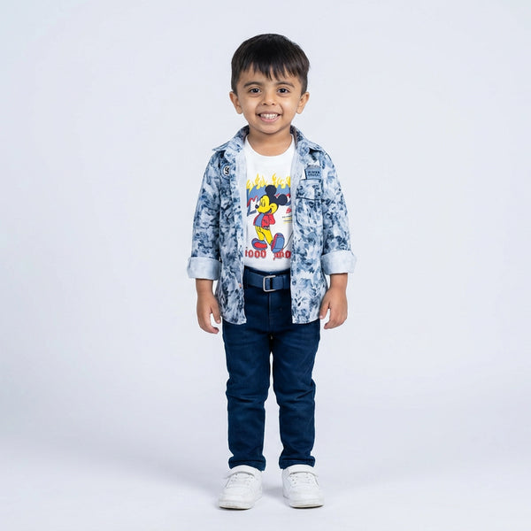 Young boy smiling in a blue printed shirt, cartoon graphic T-shirt, blue jeans, belt, and white sneakers, standing against a plain light background.