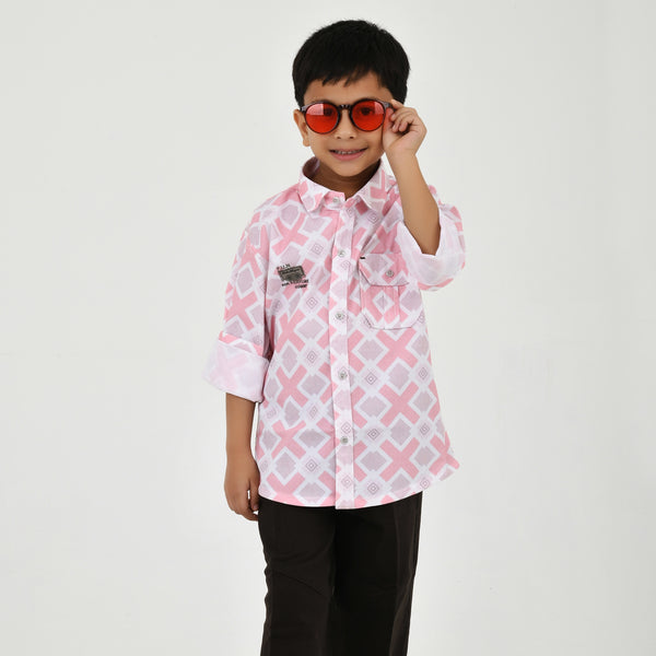 Young boy wearing a geometric print casual shirt with dark pants and white sneakers, shown in pink and green color variants against a plain light background.