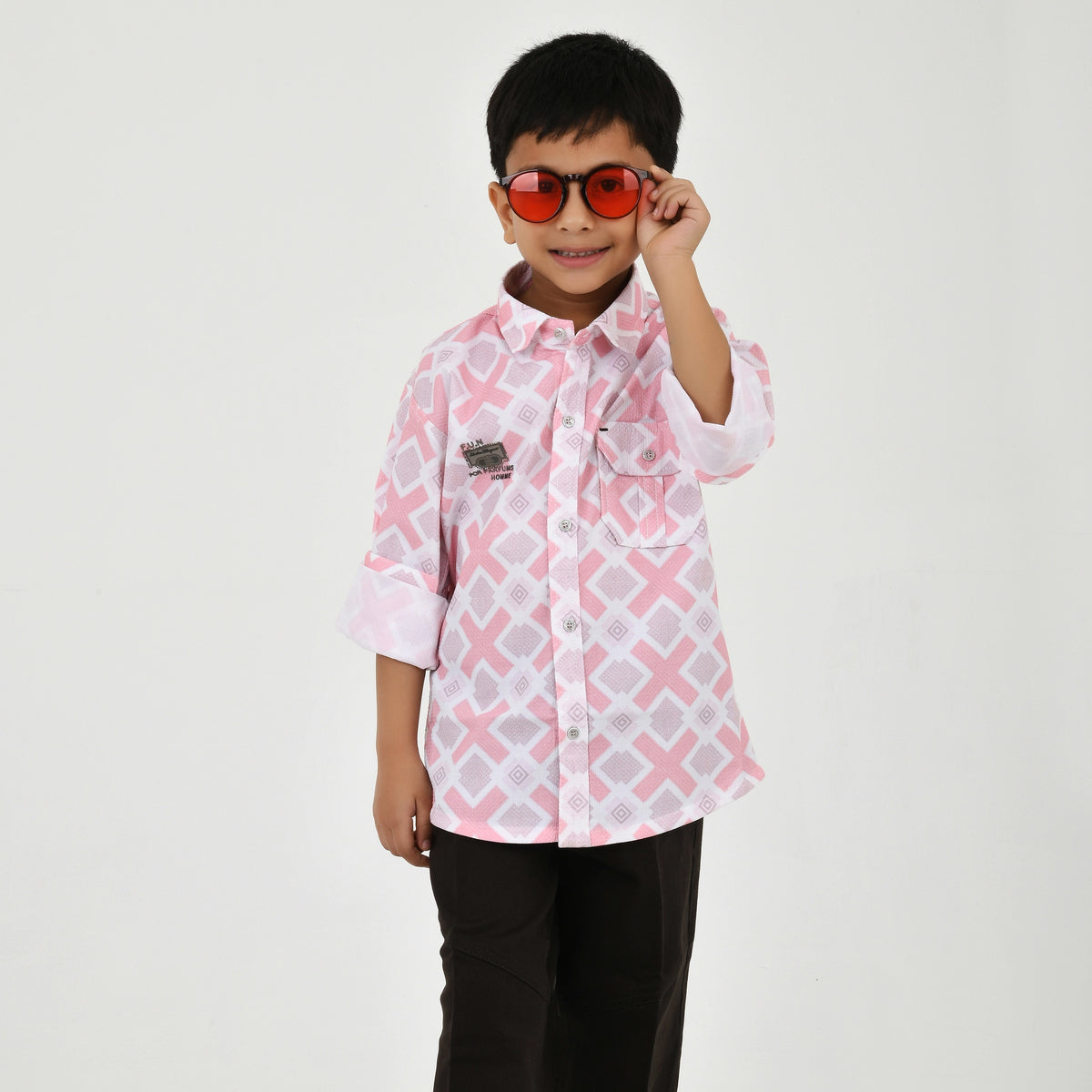 Young boy wearing a geometric print casual shirt with dark pants and white sneakers, shown in pink and green color variants against a plain light background.