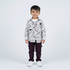 Young boy smiling in a grey abstract printed shirt over a graphic T-shirt, maroon cargo pants, and white sneakers, standing against a plain light background.