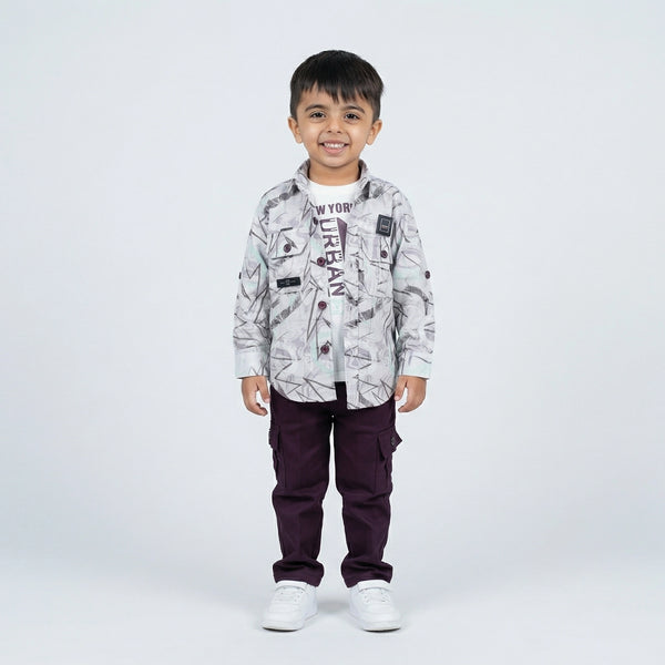 Young boy smiling in a grey abstract printed shirt over a graphic T-shirt, maroon cargo pants, and white sneakers, standing against a plain light background.
