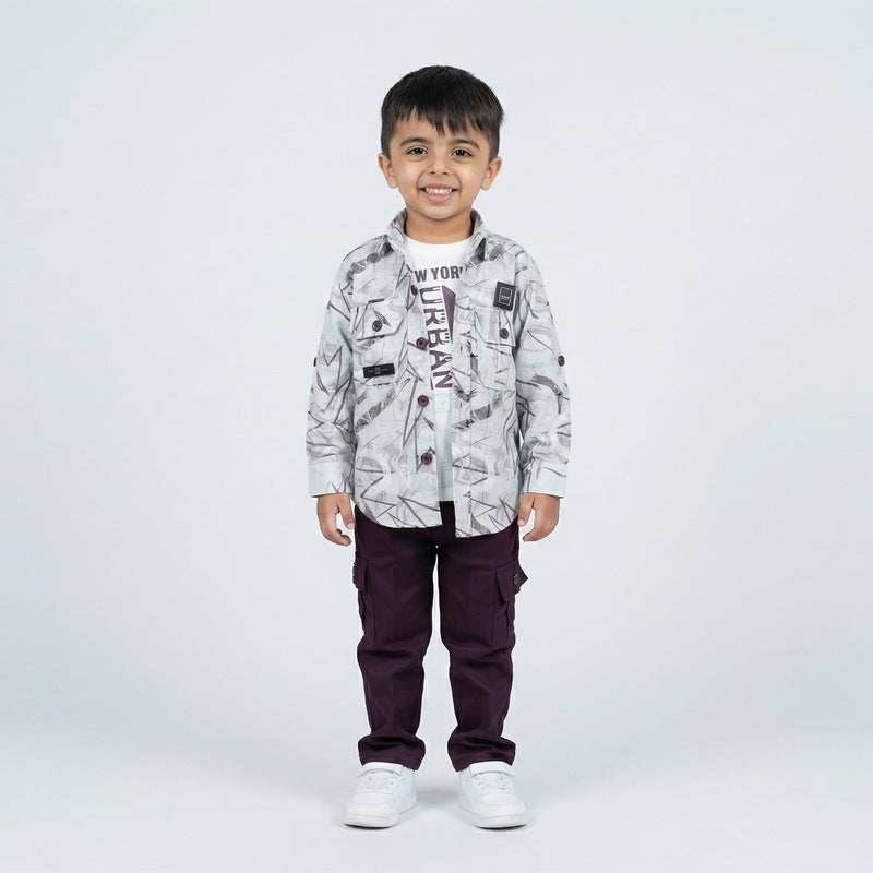 Young boy smiling in a grey abstract printed shirt over a graphic T-shirt, maroon cargo pants, and white sneakers, standing against a plain light background.