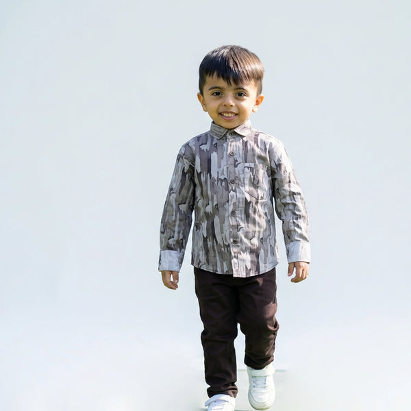 Young boy wearing a grey camouflage print full sleeve shirt, dark pants, and white sneakers, walking and smiling against a plain light background.