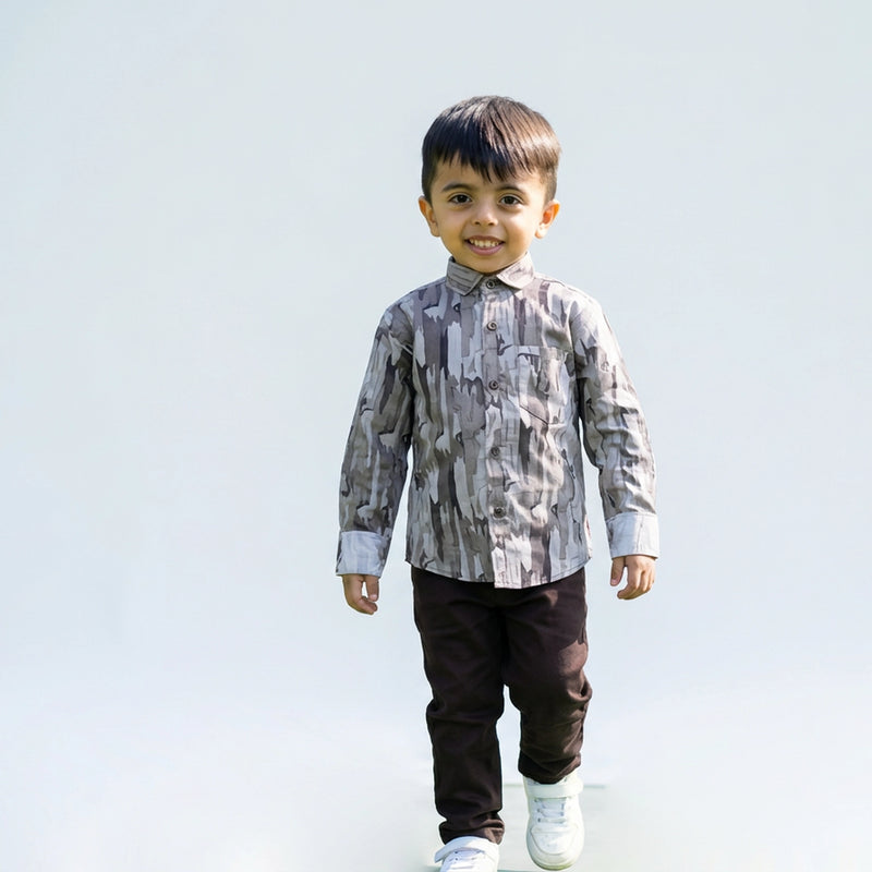 Young boy wearing a grey camouflage print full sleeve shirt, dark pants, and white sneakers, walking and smiling against a plain light background.
