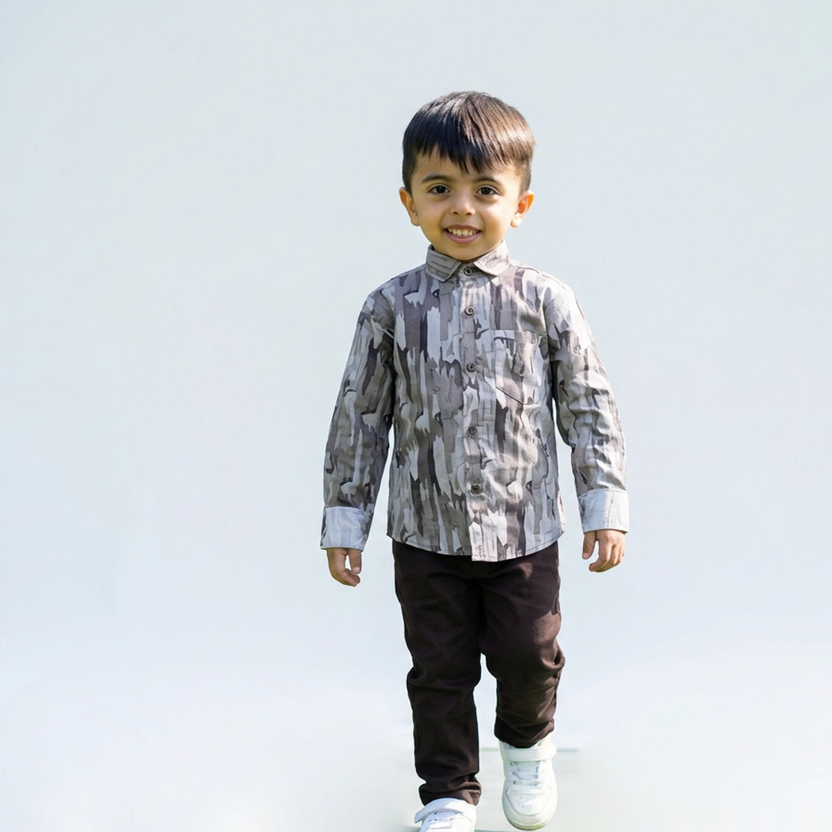 Young boy wearing a grey camouflage print full sleeve shirt, dark pants, and white sneakers, walking and smiling against a plain light background.