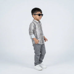 Young boy smiling in a grey printed full sleeve shirt, grey trousers, and white sneakers, standing against a plain light background.