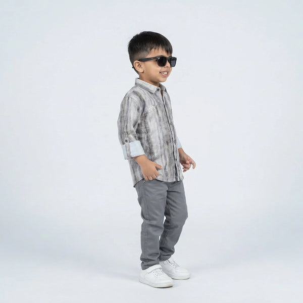 Young boy smiling in a grey printed full sleeve shirt, grey trousers, and white sneakers, standing against a plain light background.