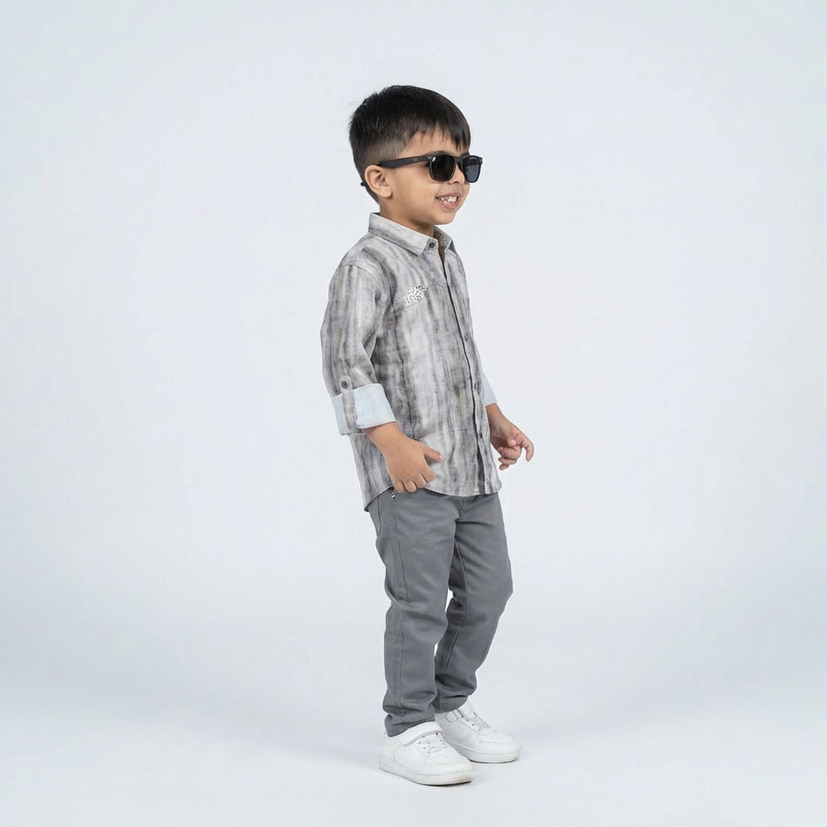 Young boy smiling in a grey printed full sleeve shirt, grey trousers, and white sneakers, standing against a plain light background.
