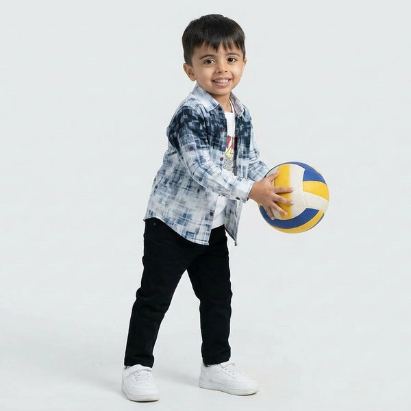 Young boy smiling in a blue printed casual shirt over a graphic T-shirt, black pants, and white sneakers, standing against a plain light background.