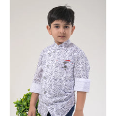 Young boy wearing a white abstract print shirt, blue jeans, and sport sneakers, posing indoors on a round rug beside a small table and plant.