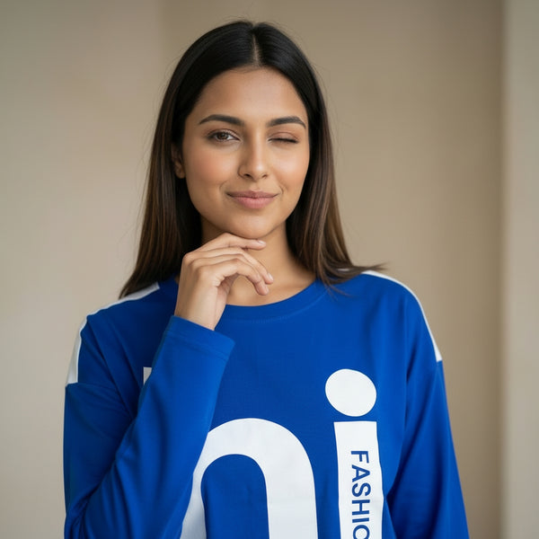 Woman wearing a blue long-sleeve top with graphic text, posing with a hand under her chin and winking against a neutral background.

