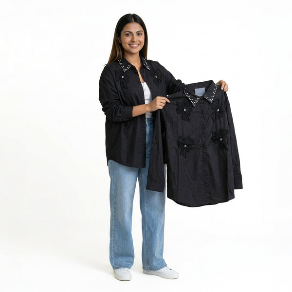 A smiling woman wearing a black button-down shirt with floral embellishments and studded collar detailing, paired with light blue jeans and white sneakers, holding a matching black shirt.