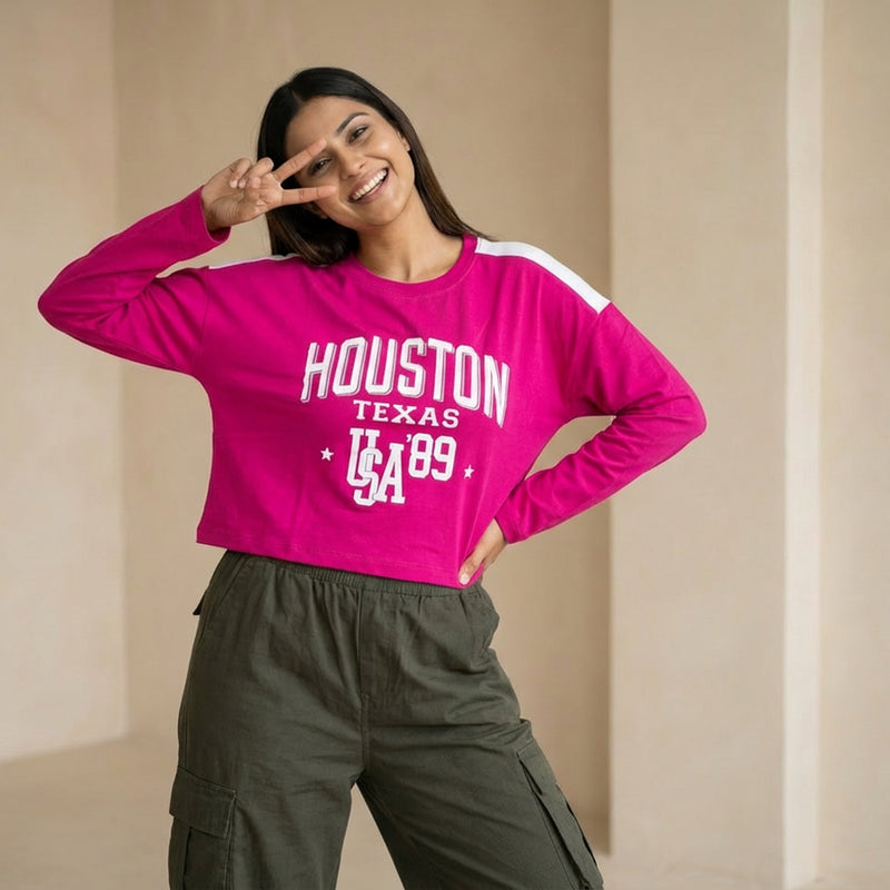 Woman wearing a bright pink cropped sweatshirt with “Houston Texas USA ’89” text, paired with olive green cargo pants, posing with a peace sign gesture.

