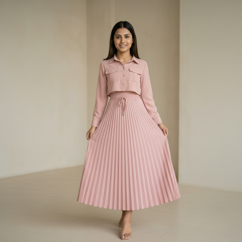 Woman wearing a dusty pink cropped shirt with front pockets paired with a matching pleated midi skirt, standing in a minimal indoor setting.

