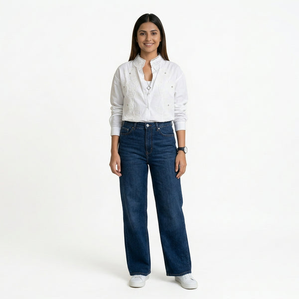 A woman wearing a white button-down shirt with delicate embroidered detailing and a textured collar, paired with blue wide-leg jeans and white sneakers.