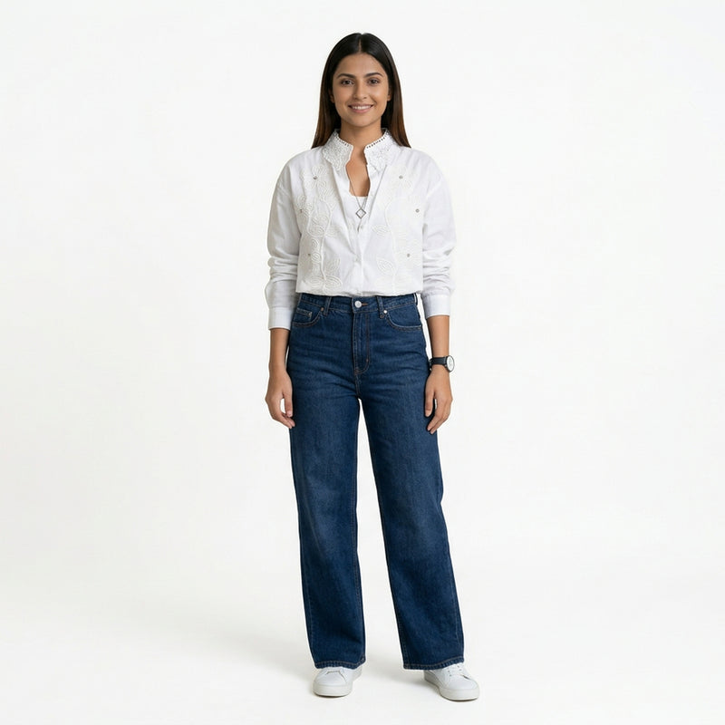 A woman wearing a white button-down shirt with delicate embroidered detailing and a textured collar, paired with blue wide-leg jeans and white sneakers.