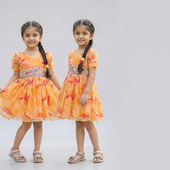 Two young girls wearing matching orange dresses with floral patterns on a plain background