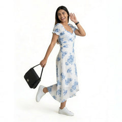 A smiling woman wearing a white midi dress with blue floral prints, paired with white sneakers. She is posing playfully on one leg, holding a black handbag, and looking over her shoulder against a plain white background.