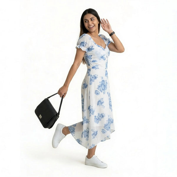 A smiling woman wearing a white midi dress with blue floral prints, paired with white sneakers. She is posing playfully on one leg, holding a black handbag, and looking over her shoulder against a plain white background.
