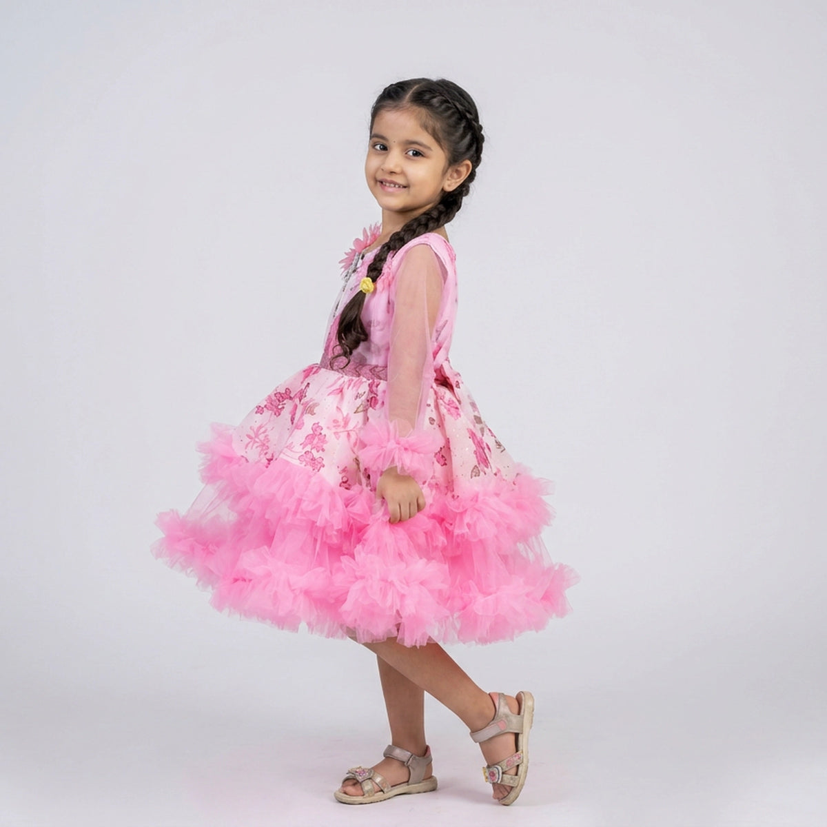 A young girl with dark braided hair is wearing a pink sleeveless dress with a sheer upper bodice, a floral patterned bodice, and a voluminous fluffy pink tulle skirt. She is wearing light brown sandals and posing against a solid light gray studio background.