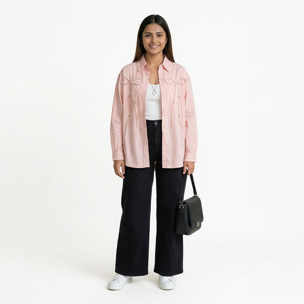 A smiling woman wearing a light pink button-down shirt with bow detailing, layered over a white top, paired with black wide-leg jeans and white sneakers, holding a structured black handbag.