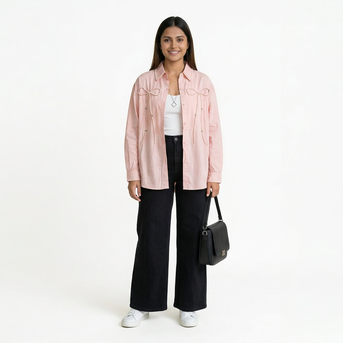 A smiling woman wearing a light pink button-down shirt with bow detailing, layered over a white top, paired with black wide-leg jeans and white sneakers, holding a structured black handbag.
