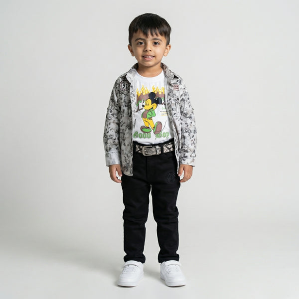 Young boy wearing a grey printed shirt over a white graphic t-shirt with black trousers, belt, and white sneakers against a light studio background